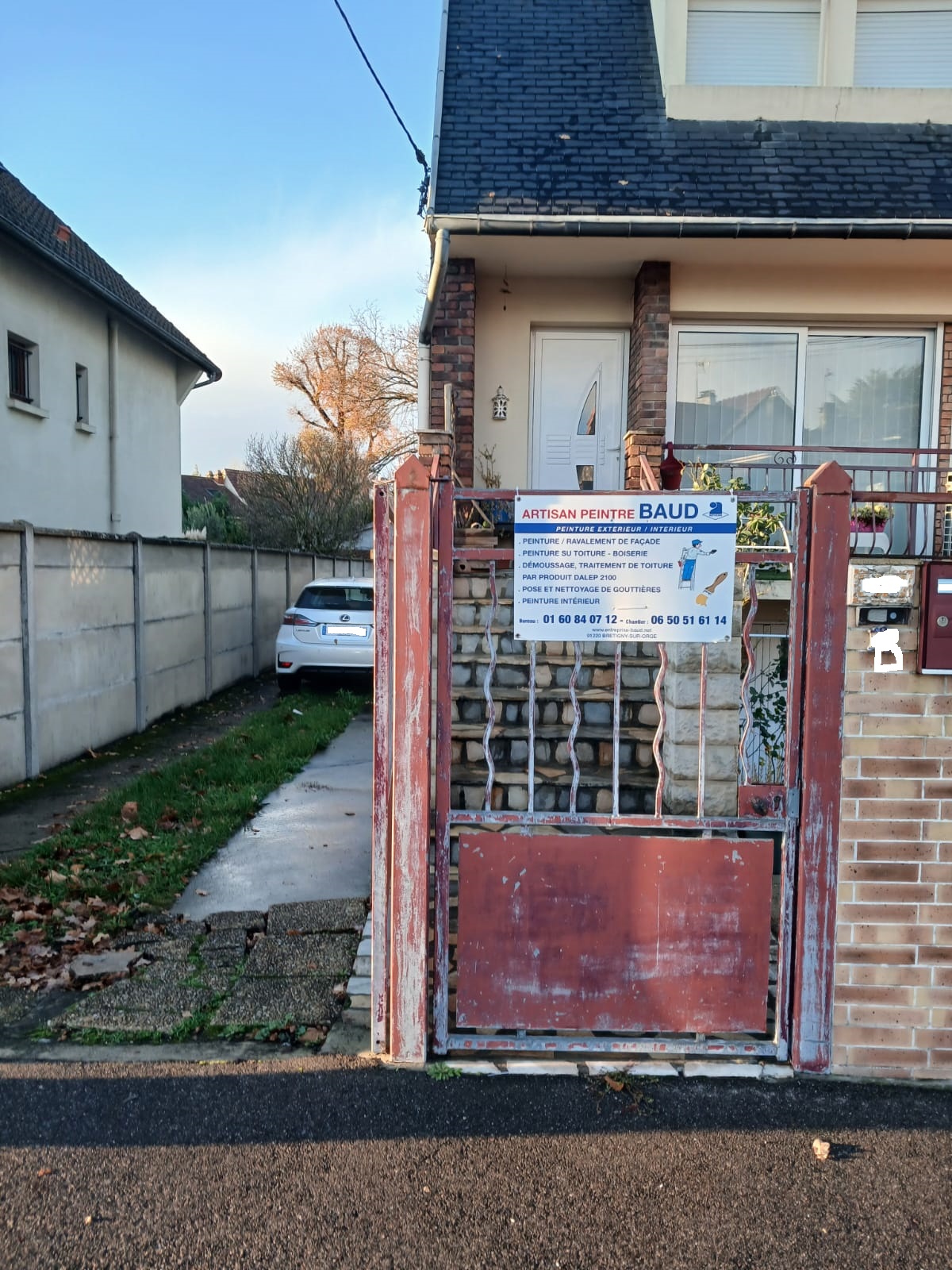 travauxà venir Entreprise BAUD Front view of a brick house with a red metal gate and a sign for a painter Artisan Peintre Baud posted on the gate A white car is parked behind the gate along a narrow driveway