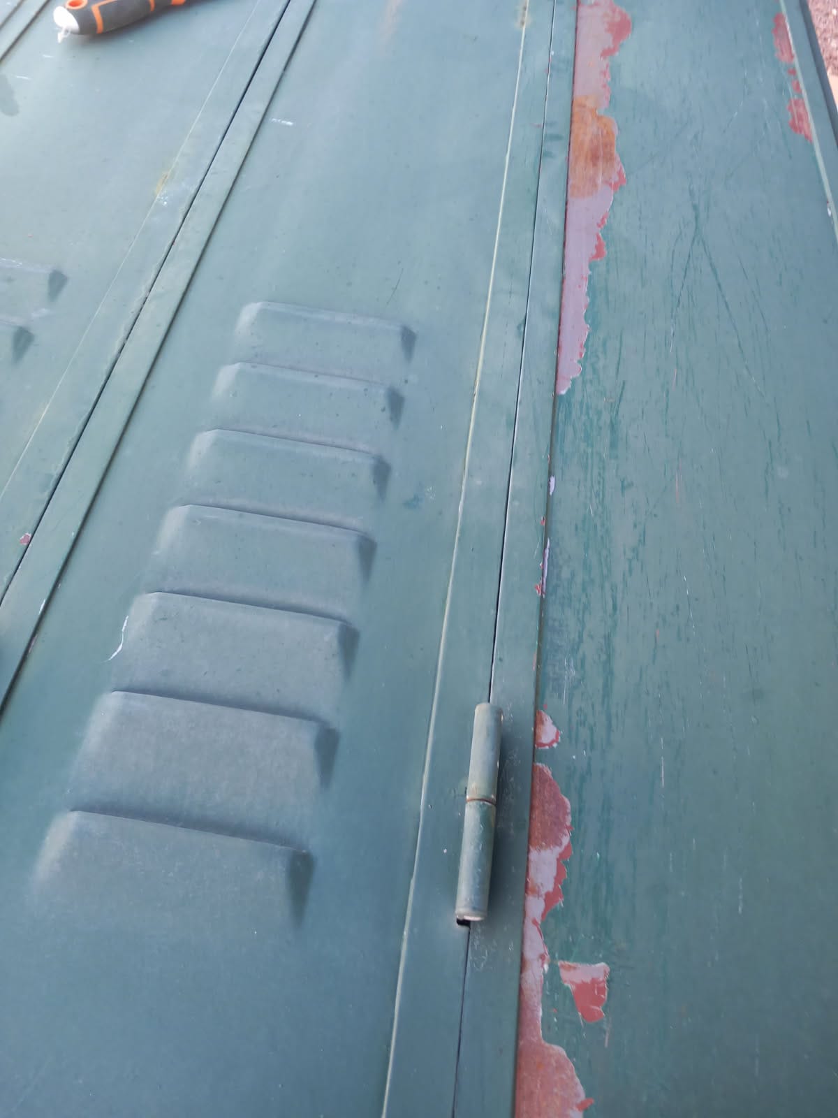 volets avant Entreprise BAUD Close up of a green metal surface with a hinge and peeling red paint along the edge plus a screwdriver in the top corner